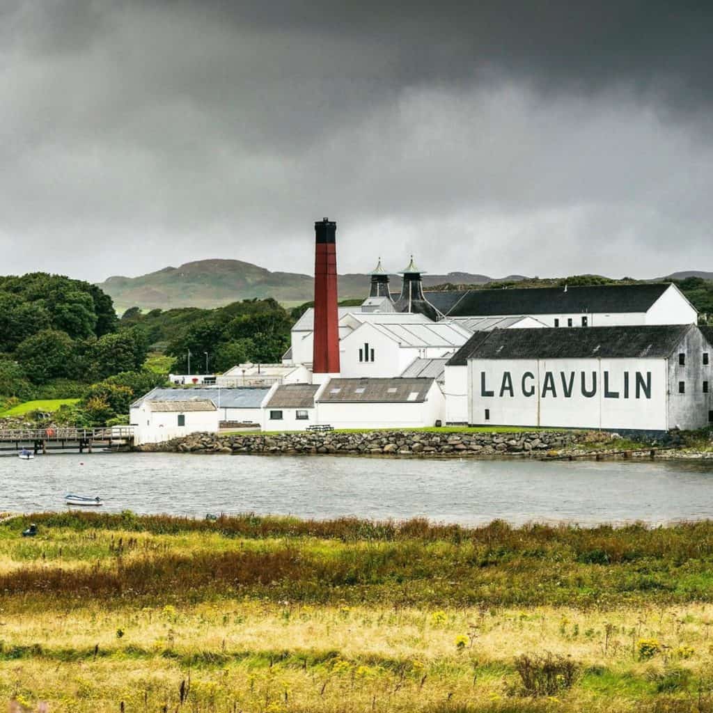 Lagavulin Distillery in picturesque countryside with lake in front and white wooden building with red chimney