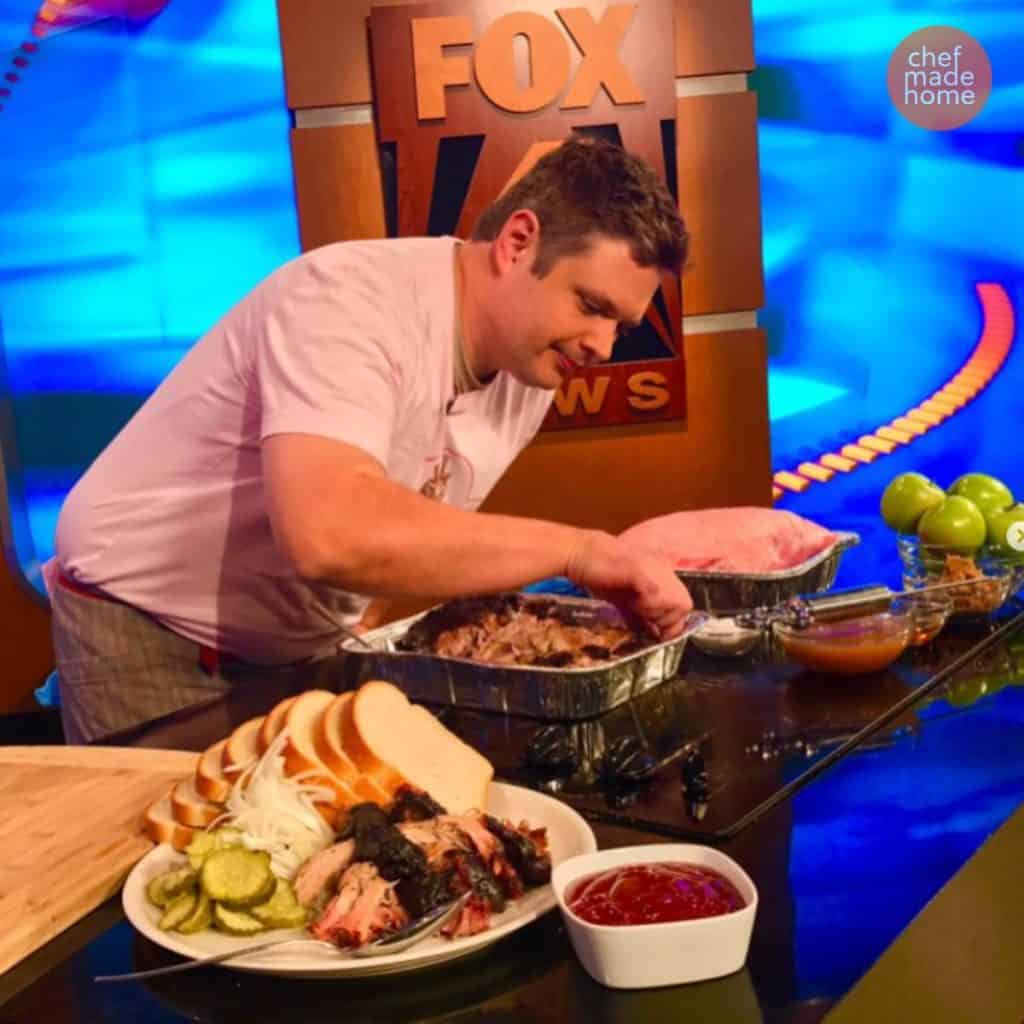 Chef Chad on a news set at Fox News Station dallas. Working on a shiny black countertop. He is picking at a piece of pulled pork in an aluminum pan with a plate of the pulled pork on a plate in the foreground. A raw pork butt is in the background next to a meat syringe and a orange sauce.